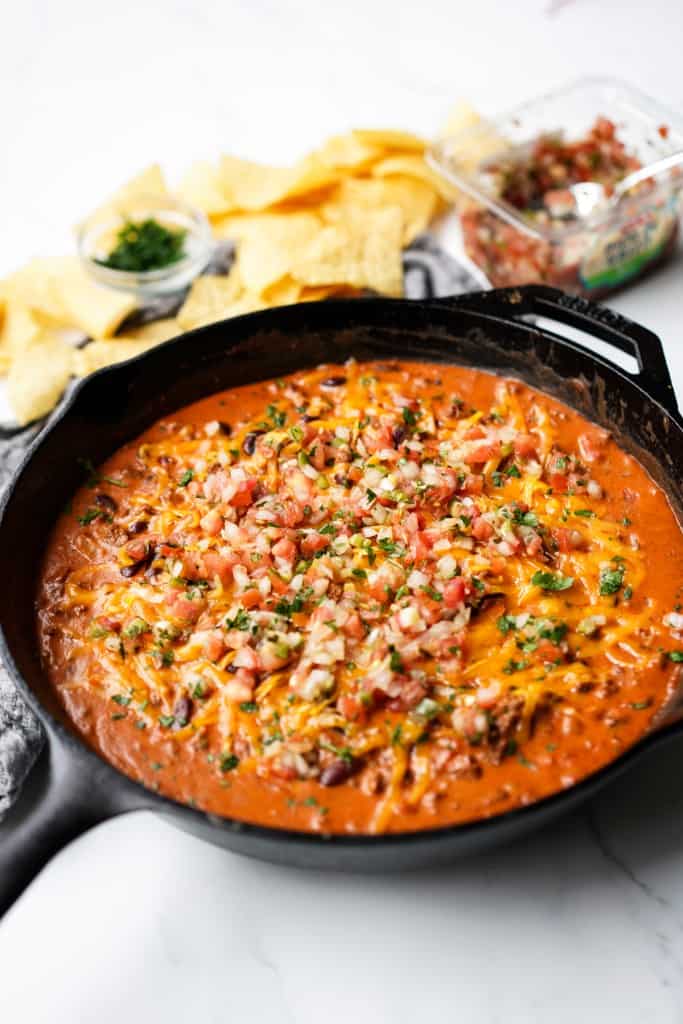 A black cast iron skillet is filled with a chili cheese dip that is topped with cheddar cheese and pico de gallo.