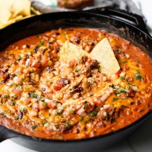 A cast iron skillet filled with beans, cheese, and ground beef topped with cheese and pico de gallo and chips ready to be eaten.