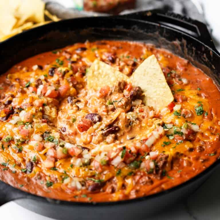 A cast iron skillet filled with beans, cheese, and ground beef topped with cheese and pico de gallo and chips ready to be eaten.