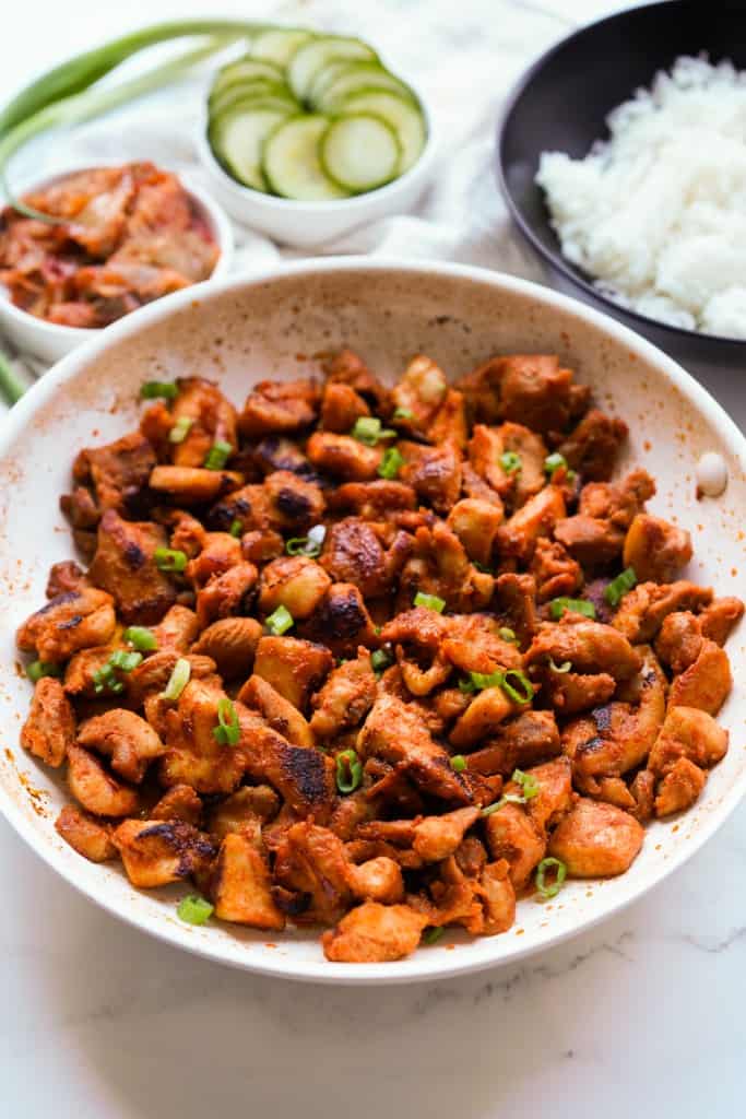 A skillet with pieces of chicken thighs seasoned with gochujang, with kimchi and cucumbers in the background