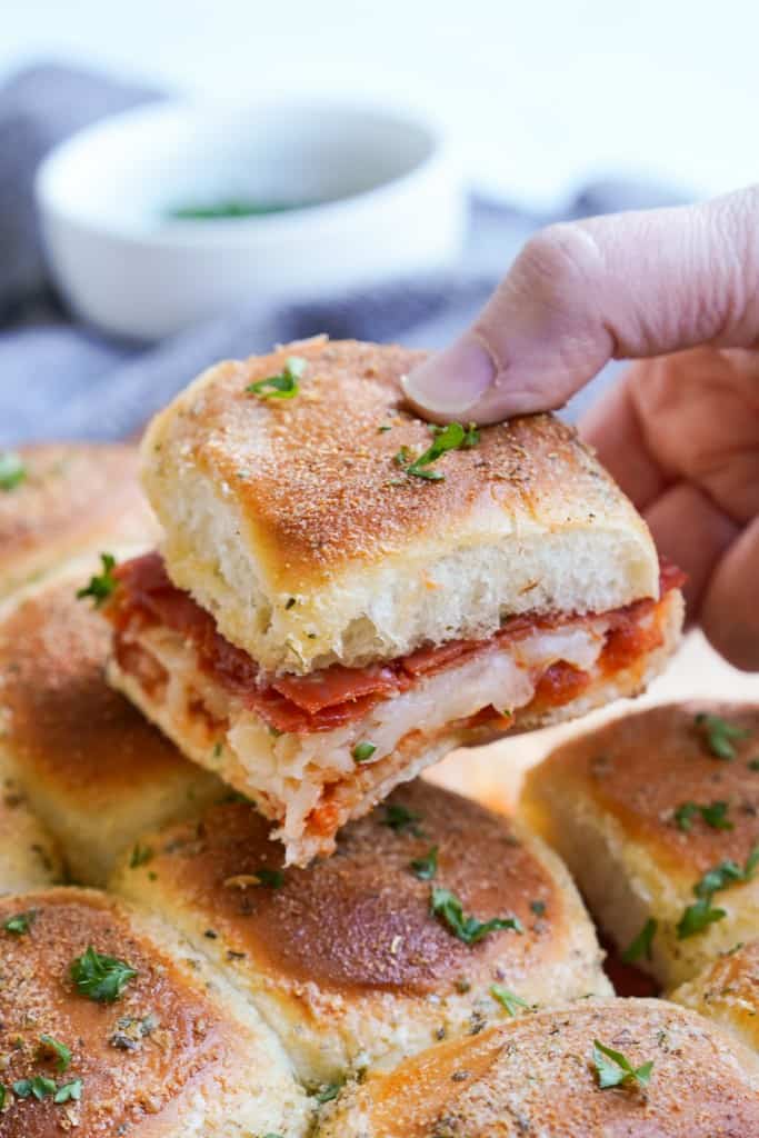 A hand is holding one pepperoni pizza slider stuffed with gooey cheese and pepperoni just over the other sliders.