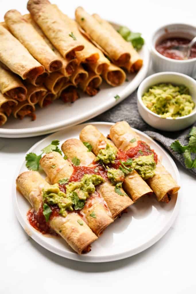 A plate of homemade chicken taquitos topped with salsa, guacamole, and cheese dip.