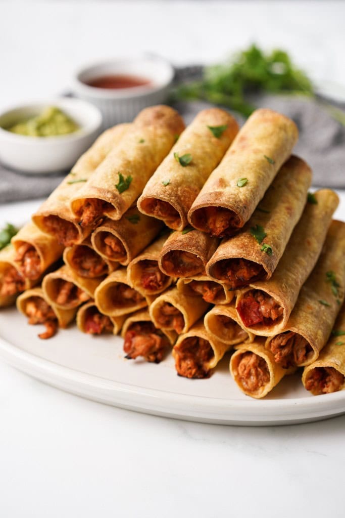 A huge stack of crispy baked chicken taquitos on a white platter.