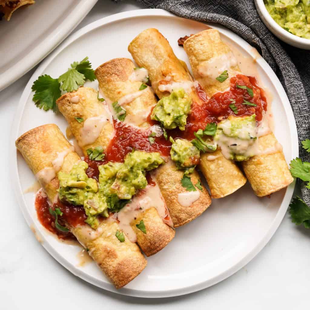 A plate of five homemade chicken taquitos topped with sauces and ready to eat.