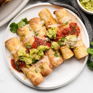 A plate of five homemade chicken taquitos topped with sauces and ready to eat.