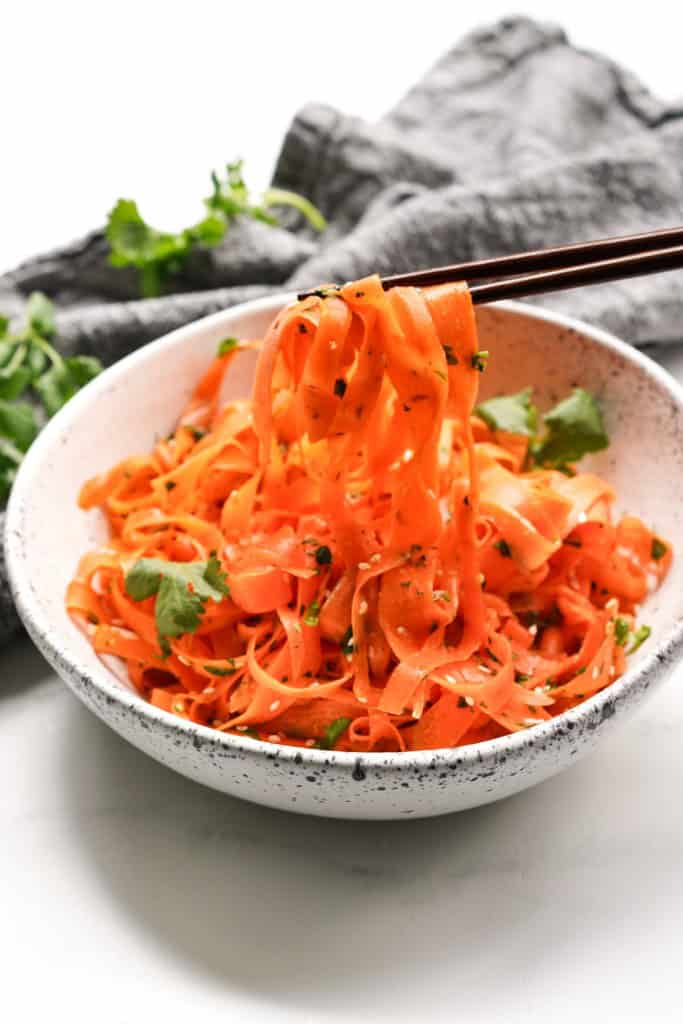 A pair of chopsticks are holding some Asian ribbon salad over a bowl.