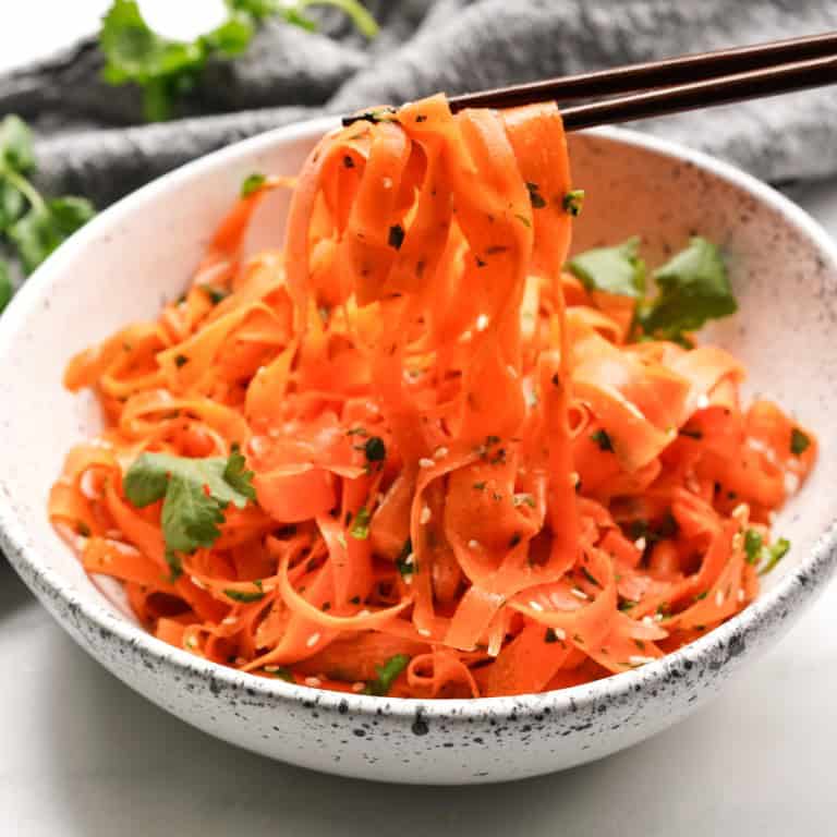 A close-up picture of chopsticks picking up some Asian carrot ribbon salad out of a bowl.