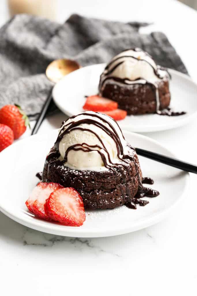 Two chocolate lave cakes are on individual small plates topped with ice cream with sliced strawberries placed next to them.