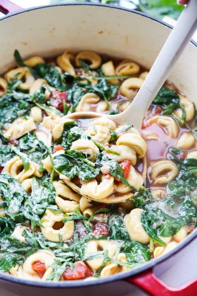 A ladle full of tortellini, spinach, tomatoes, white beans, and broth is being lifted out of a large Dutch oven.