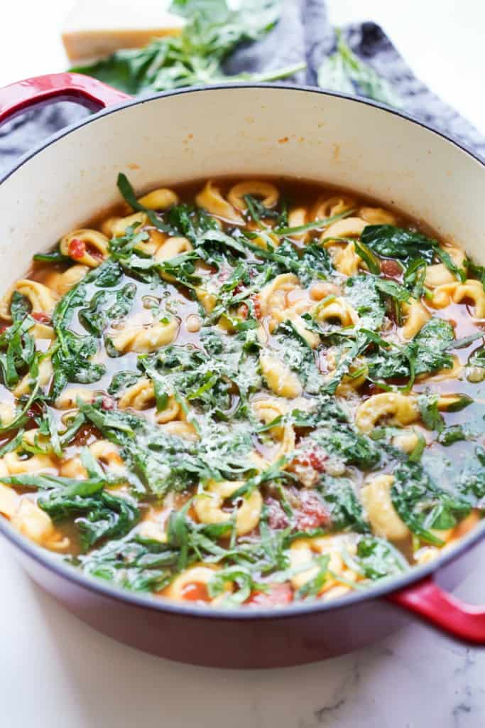 A large Dutch oven is filled with Tortellini Soup with spinach, tomatoes, topped with grated parmesan.