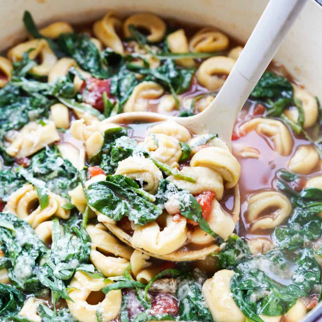 A close up picture of a ladle full of Spinach Tortellini Soup over a pot full of tortellini, tomatoes, spinach, basil, white beans, and flavorful broth.
