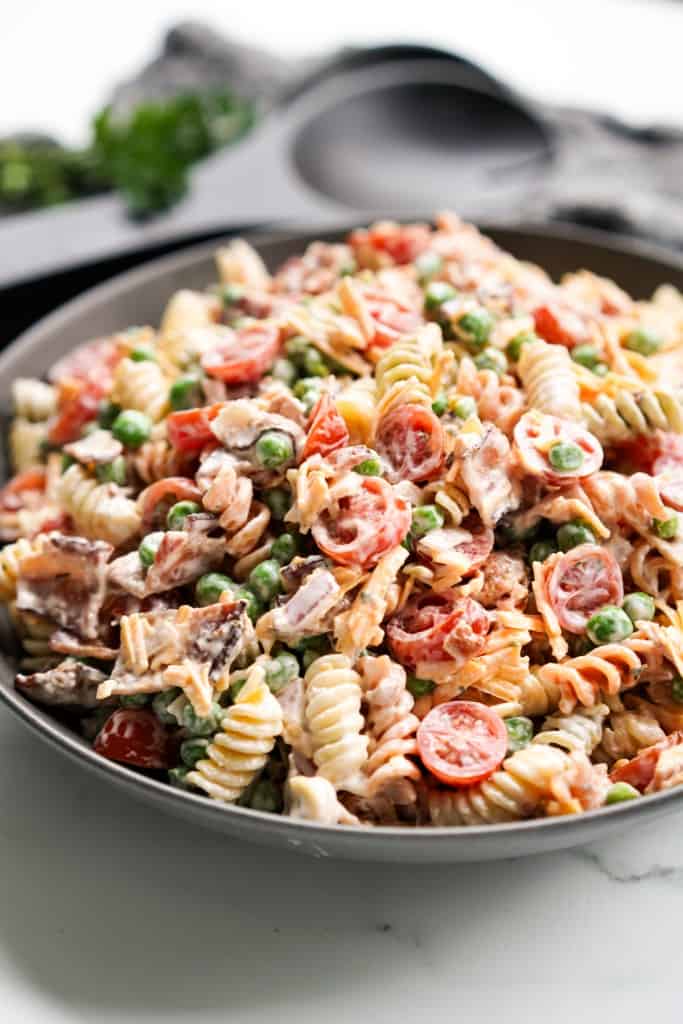 A bowl is filled with Bacon ranch cheddar pasta salad featuring sliced cherry tomatoes, peas, red onions, and bacon.