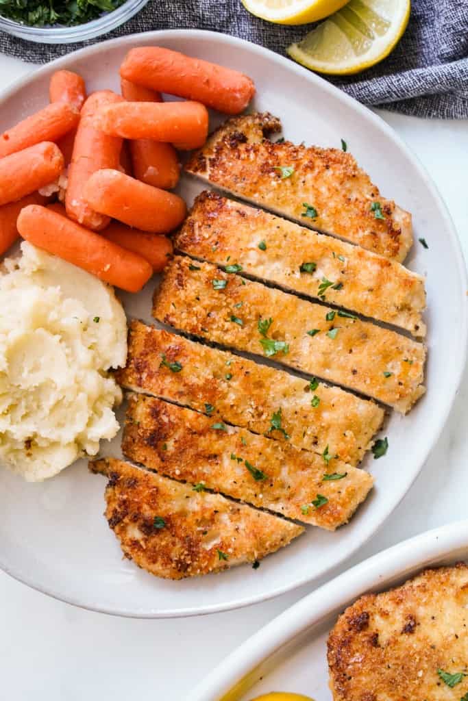 Sliced baked chicken cutlets breaded in breadcrumbs on a plater with mashed potatoes and baby carrots