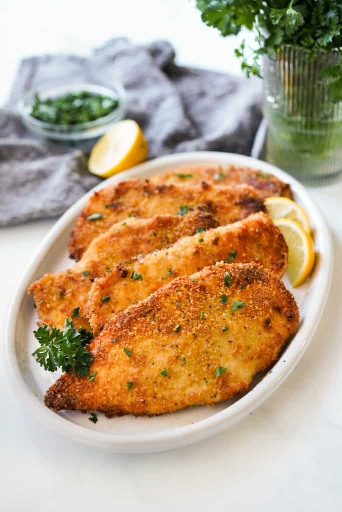 Crispy baked chicken cutlets on an oval platter with lemon wedges and fresh herbs on the side. 