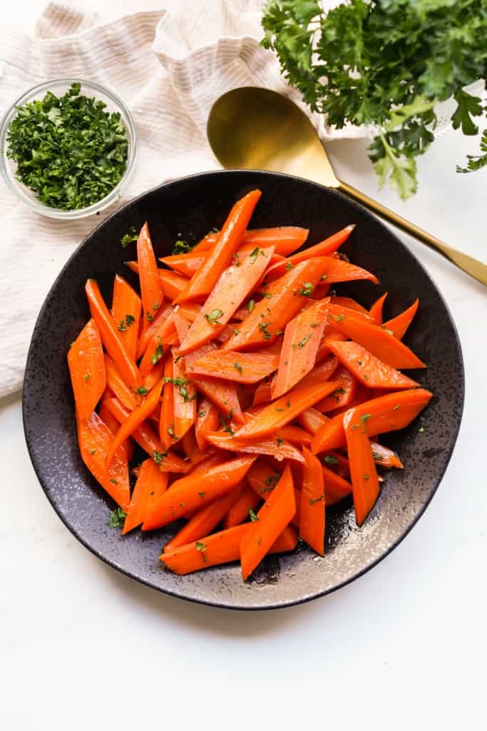 Top down view of a black bowl of candied carrots with herbs in the background.