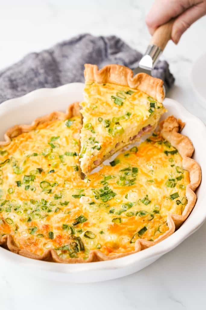 A slice of Ham and Cheese Quiche stuffed full of ham, cheese, green onions, and eggs is being lifted out of a pie pan.