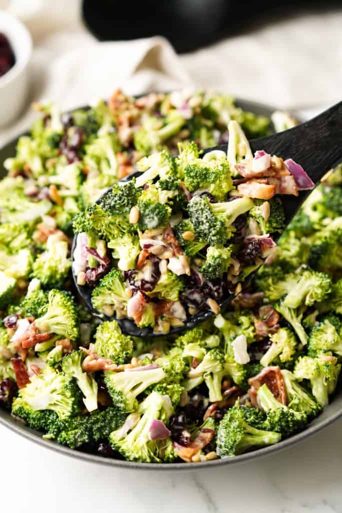 A scoop of Cold Broccoli Bacon Salad with broccoli, bacon, sunflower seeds, dried cranberries, and red onion topped with a delicious dressing is being lifted out of a bowl.