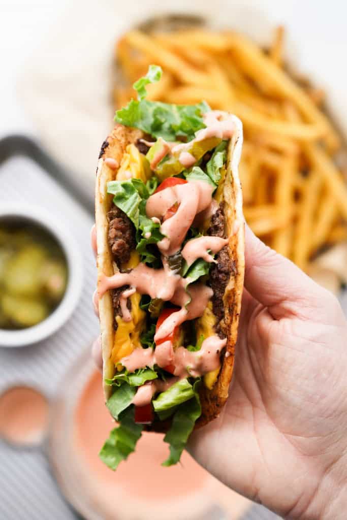 A hand is hold a Smash Burger Taco loaded with season beef, cheese, lettuce, tomato, pickles, and burger sauce.