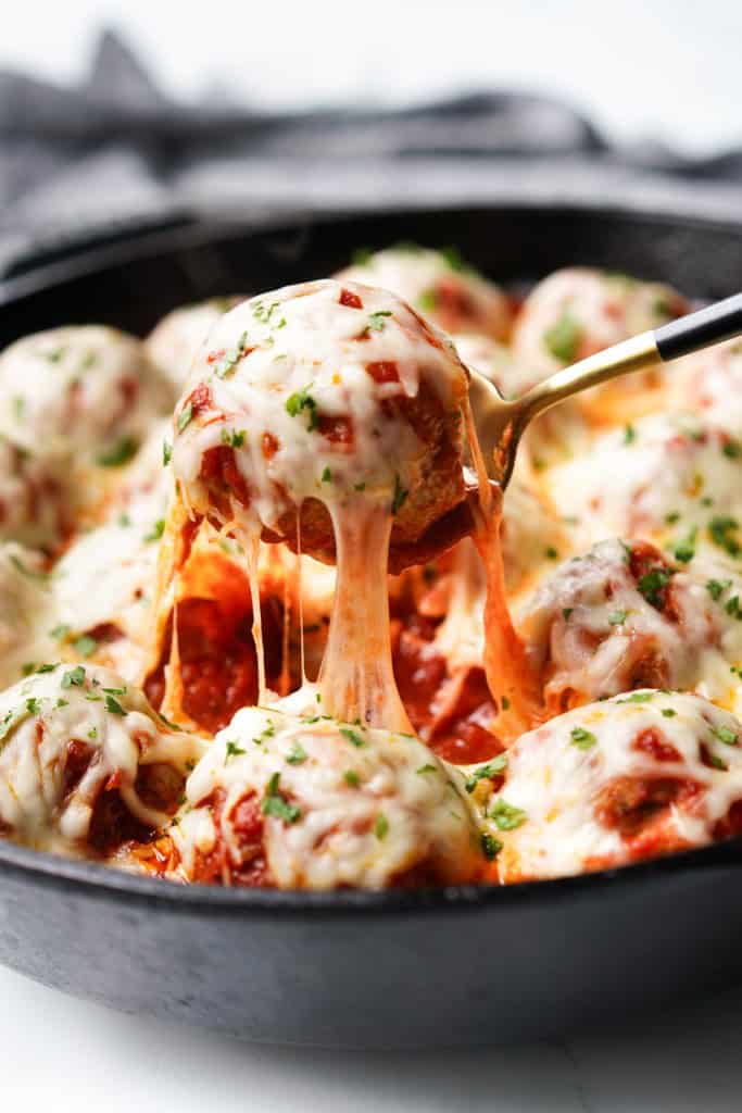 Close up picture of a chicken parm meatball being lifted out of a cast iron skillet with a yummy cheese pull.