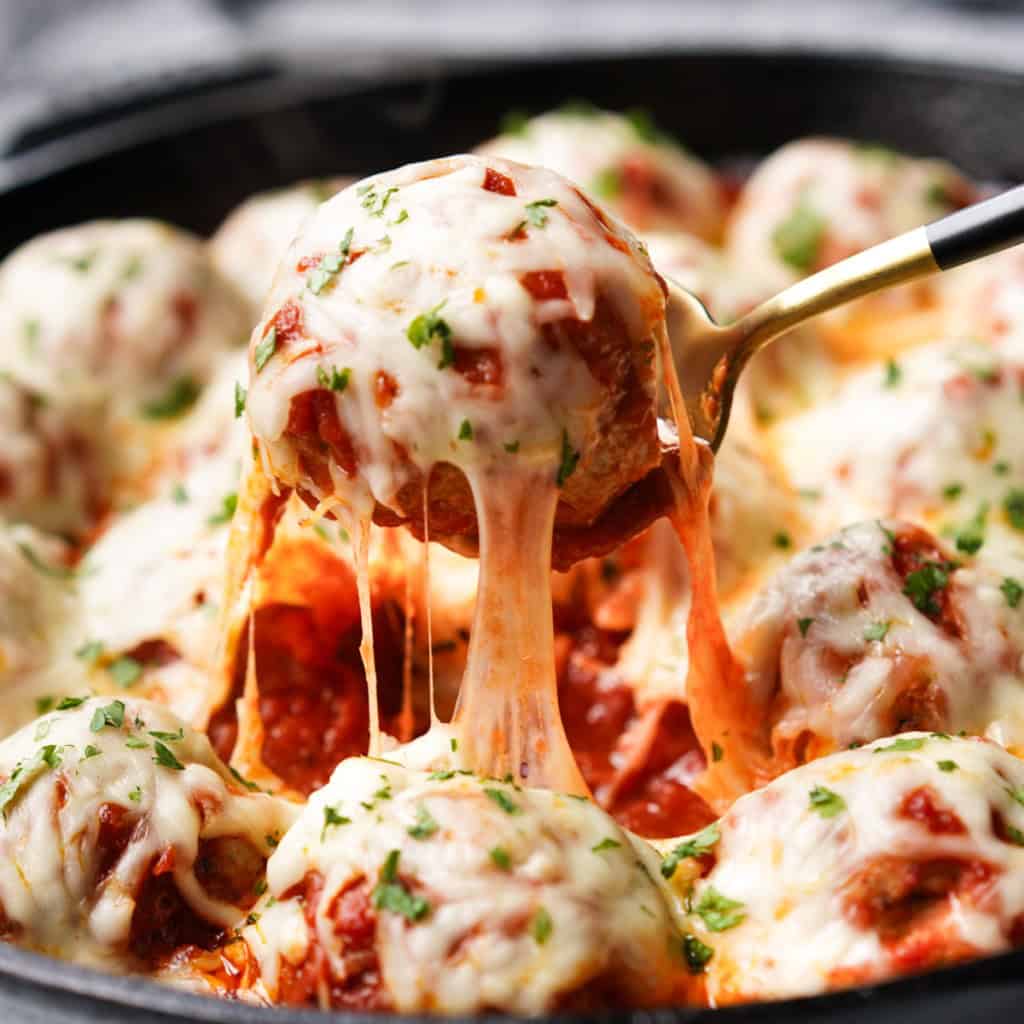 A spoon is lifting a chicken parmesan meatball out of a skillet showcasing a lovely cheese pull.