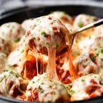 A spoon is lifting a chicken parmesan meatball out of a skillet showcasing a lovely cheese pull.