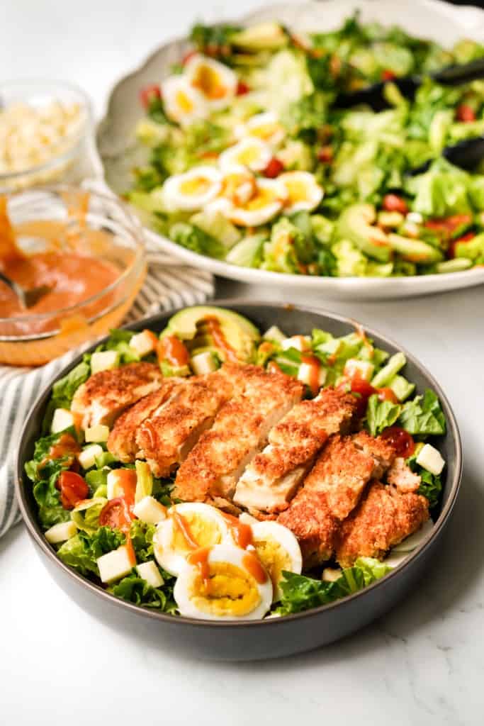 A serving of crispy chicken salad with a large platter in the background, along with honey mustard bbq dressing