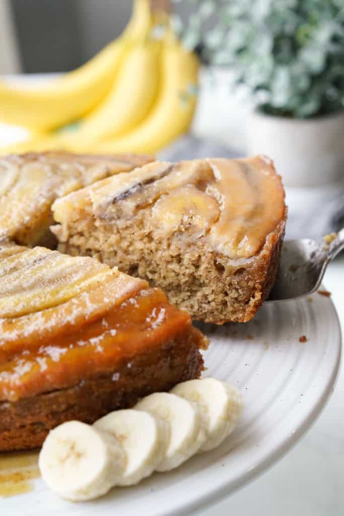 A slice of Banana Upside Down Cake has been sliced and is being lifted from the cake plate.