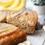 A slice of Banana Upside Down Cake topped with caramelized bananas is being removed from a cake plate.