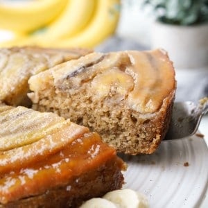 A slice of Banana Upside Down Cake topped with caramelized bananas is being removed from a cake plate.