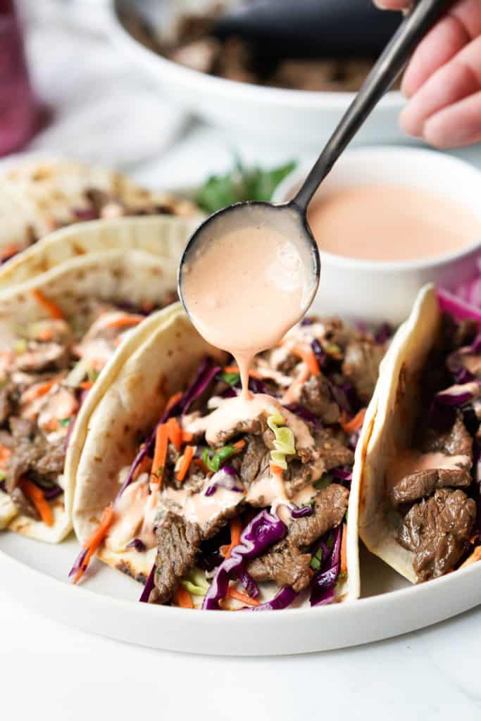 A spoon is drizzling sriracha mayo on a Korean BBQ Taco.