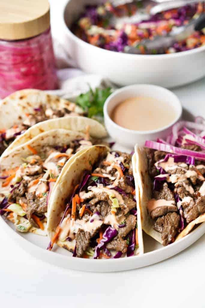 A plate is filled with four Korean street tacos with a small bowl of sriracha mayo, pickled onions, and a colorful slaw.