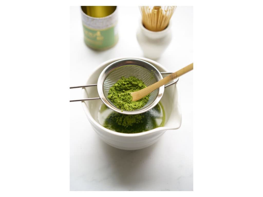 Matcha powder is being pressed through a sieve into a ceramic bowl.