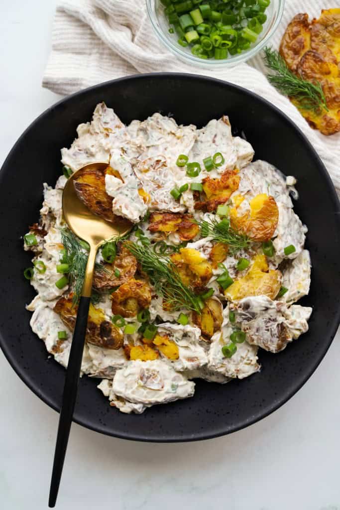A gold spoon is resting in a bowl of Crispy Smashed Potato Salad.