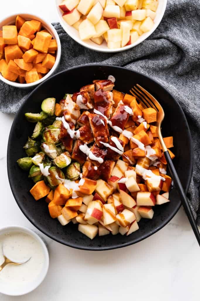 A black bowl is filled with sweet potatoes, brussels sprouts, apples, and BBQ chicken drizzled with Ranch dressing.