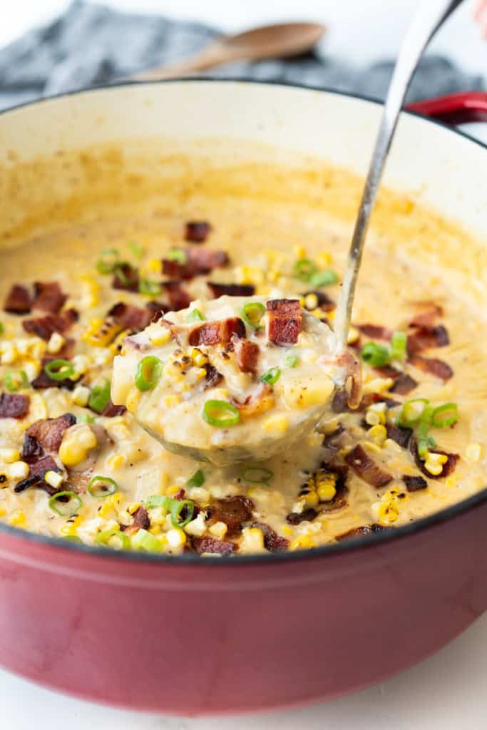 A ladle is lifting up a scoop of corn chowder with potatoes, green onion, and diced bacon.