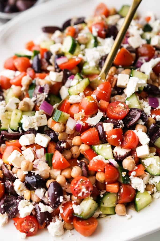 A spoon is scooping up Greek Chickpea Salad from a platter.