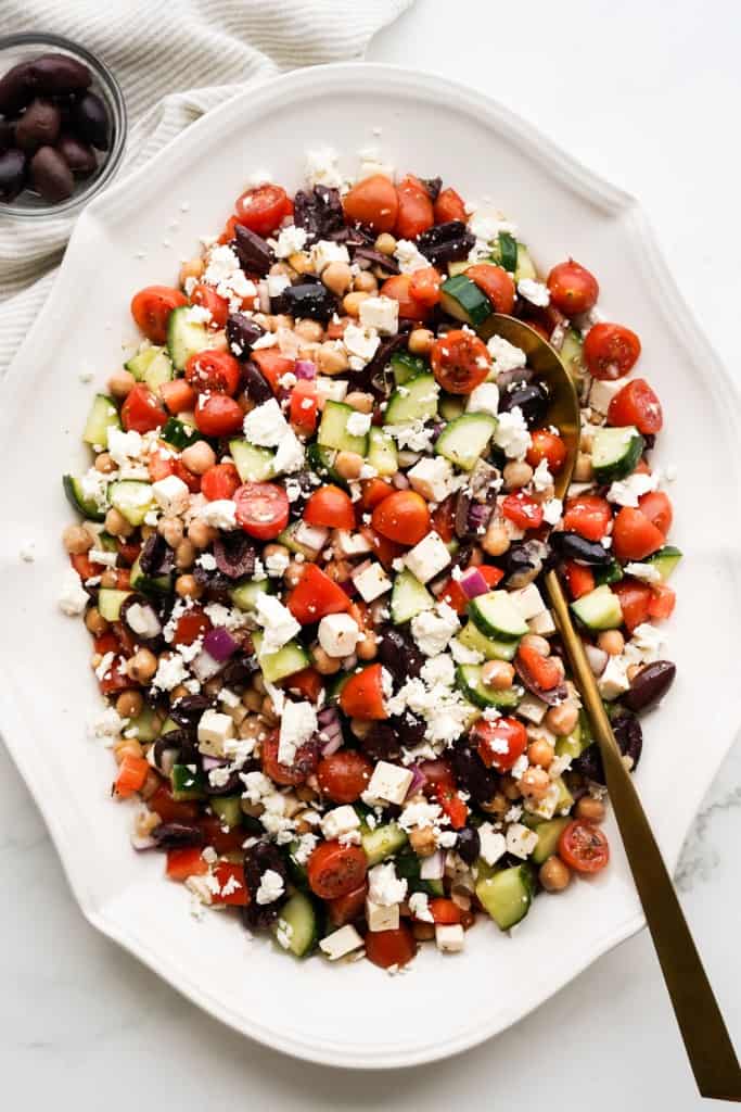 A serving spoon is resting in a platter filled with Greek salad with chickpeas.