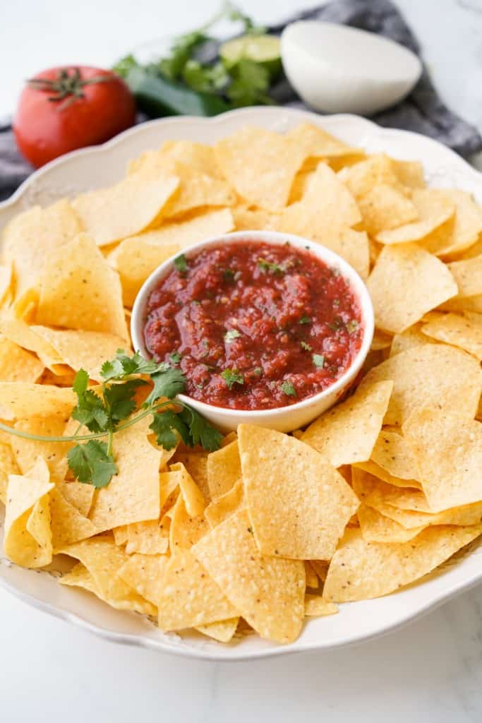 A plate of tortilla chips is surrounding a bowl of freshly made red salsa.