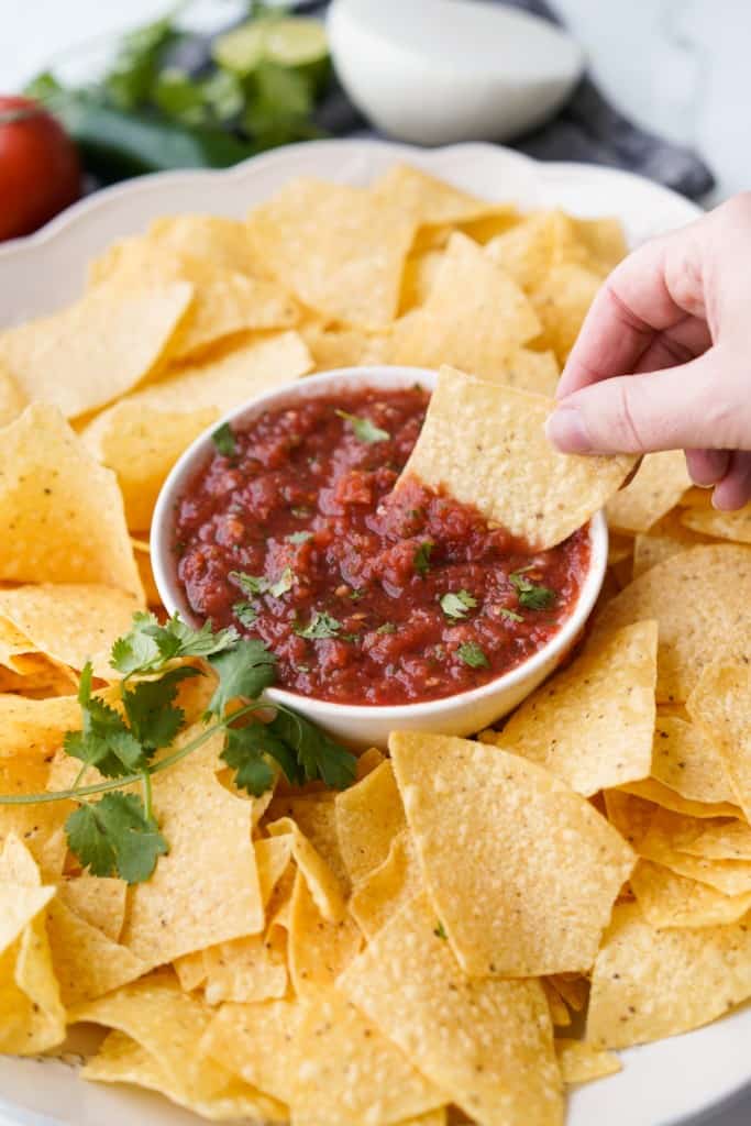 A chip is scooping tomato salsa out of a small bowl surrounded with tortilla chips.