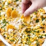 A corn chip is scooping elote dip out of a baking dish.