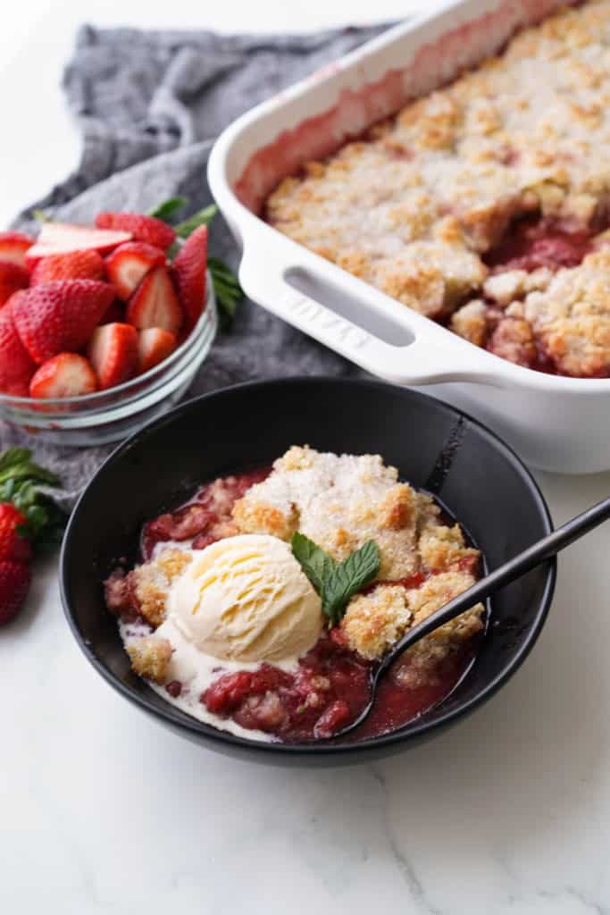 A bowl of strawberry cobbler topped with vanilla ice cream with the large serving dish and strawberries in the background.