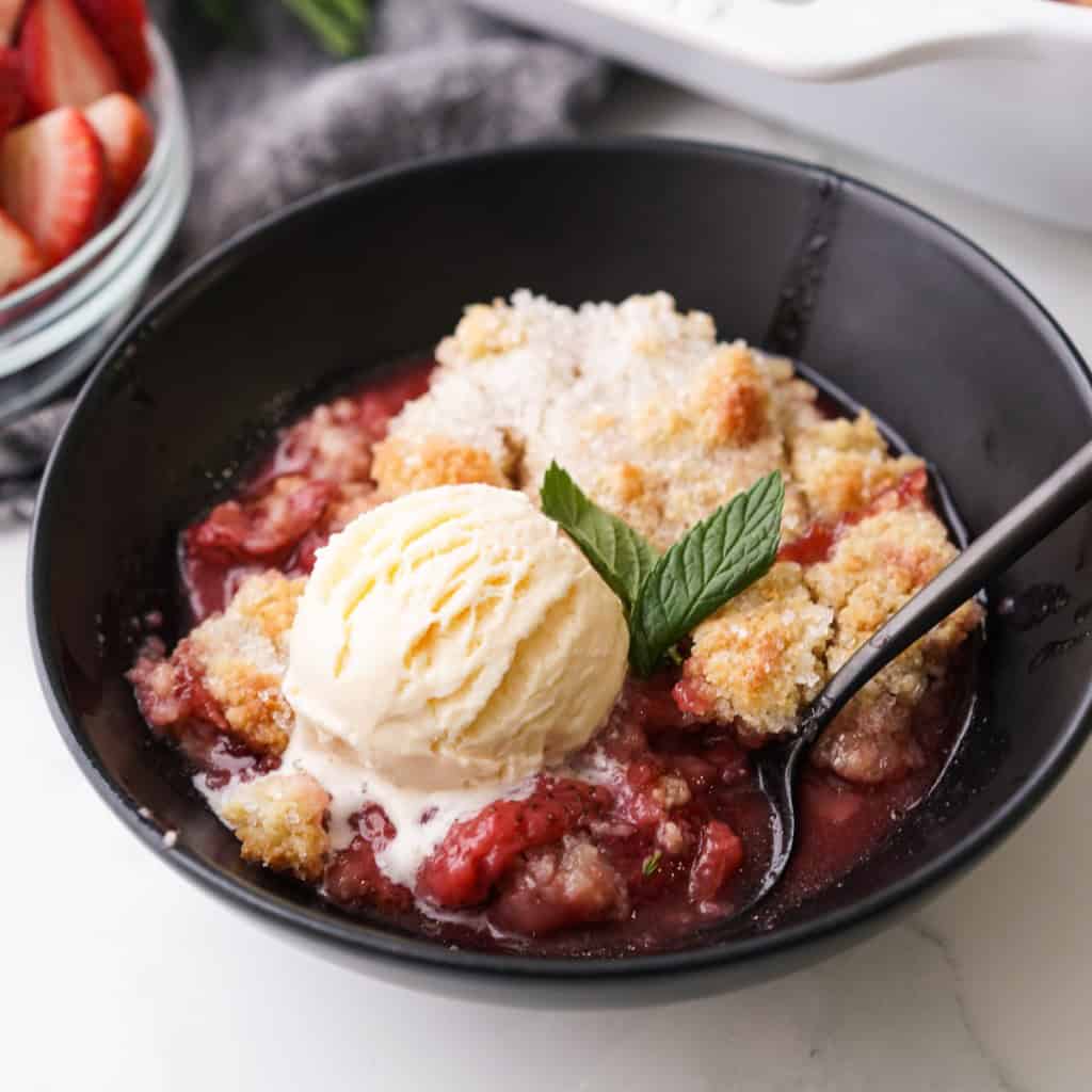 A black bowl is filled with fresh strawberry cobbler topped with vanilla ice cream.