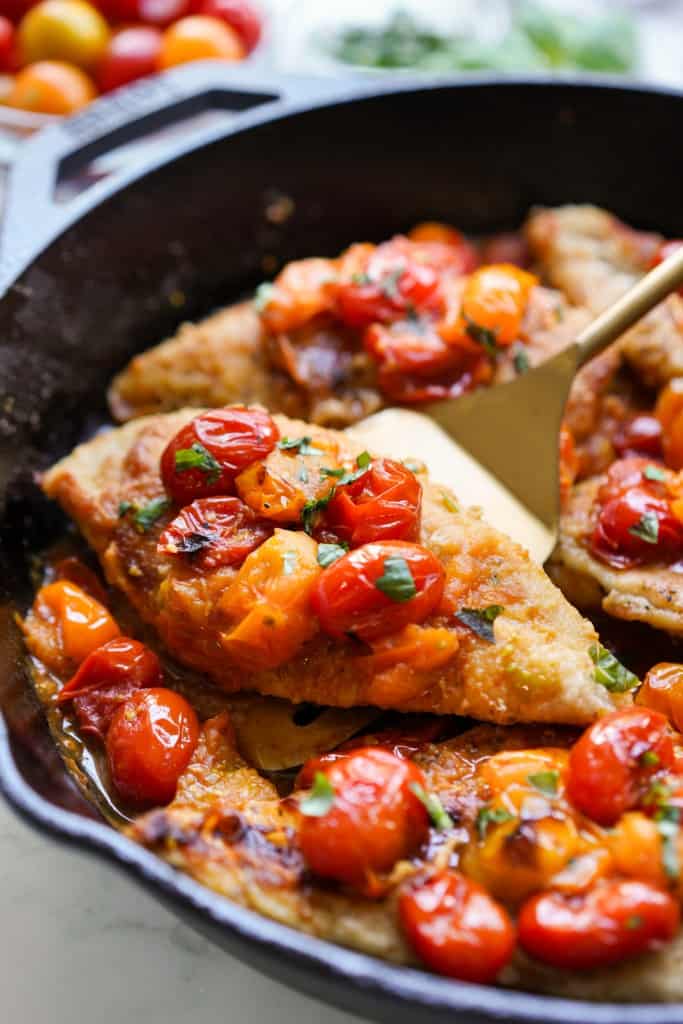 A gold spatula is lifting a chicken breast covered with blistered tomatoes.
