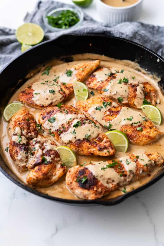 A cast iron skillet is filled with creamy coconut braised chicken garnished with lime wedges and parsley.