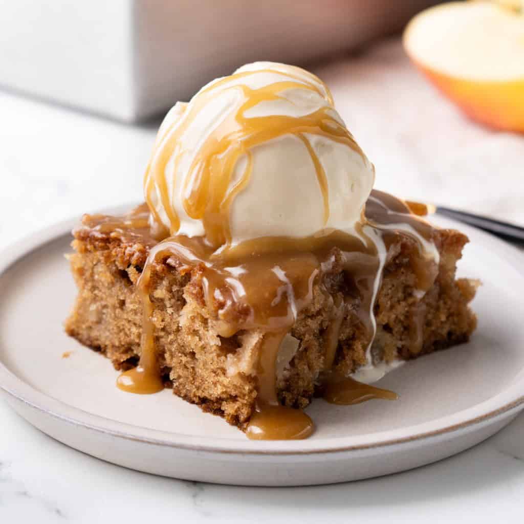 Caramel sauce is dripping down a scoop of ice cream sitting on top of a slice of caramel apple cake.