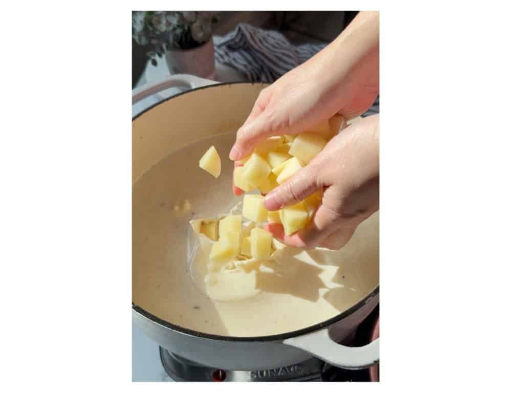 Hands adding a handful of diced raw potatoes into a Dutch oven filled with a creamy liquid base.