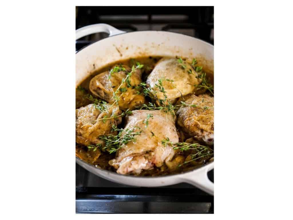 Braised chicken thighs, skin side up, in a white braiser with the wine and broth sauce, topped with fresh thyme sprigs before baking.