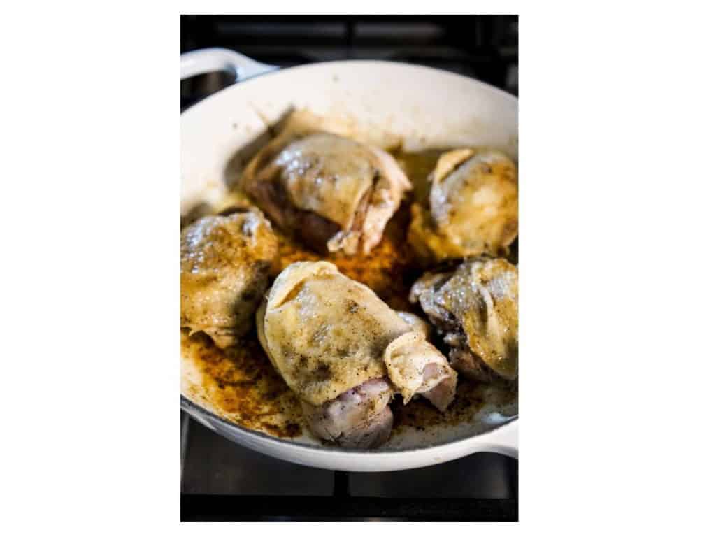 Browned bone-in, skin-on chicken thighs in a white braiser on the stovetop, with golden crispy skin and brown bits on the bottom of the pan.